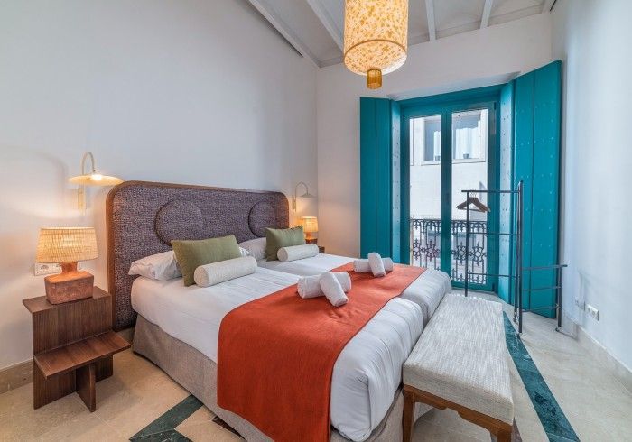 Habitación luminosa con cama, manta naranja, puertas de balcón turquesa y lámpara colgante tejida.