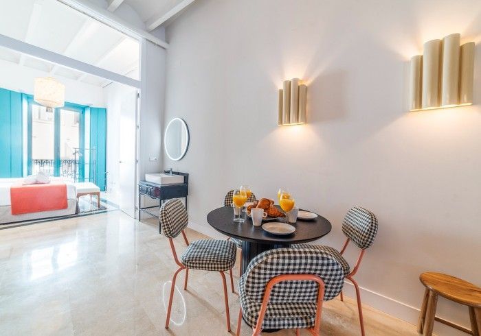 Comedor moderno y luminoso con mesa redonda, sillas estampadas, apliques de pared y centro de mesa con frutas.