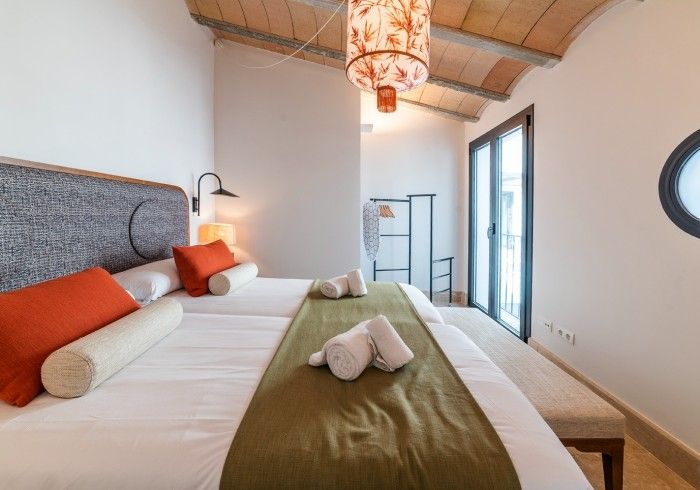Habitación luminosa con cama, cabecero gris, almohadas naranjas, manta verde y puertas que dan al balcón.