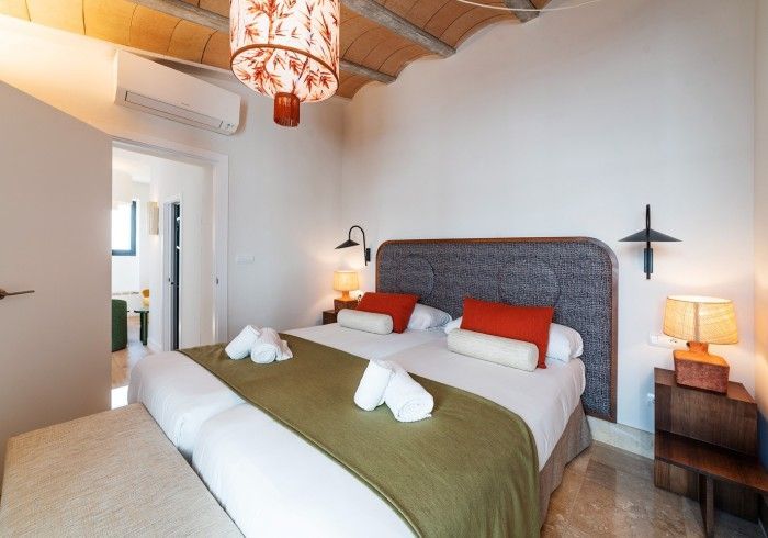 Habitación acogedora con una cama grande, manta verde, almohadas naranjas y lámparas colgantes cálidas.