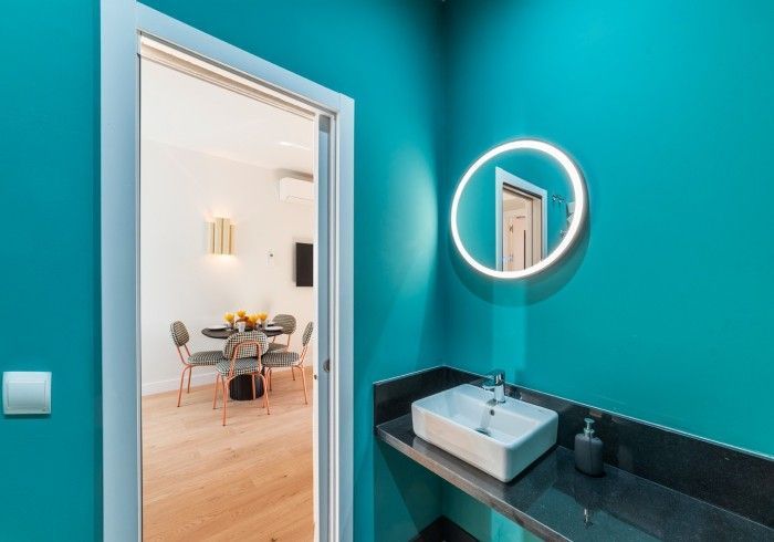 Baño color turquesa con tocador negro, espejo redondo y puerta abierta a un luminoso comedor.