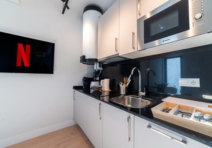 Rincón de cocina moderno con armarios blancos, salpicadero negro, fregadero, cafetera, microondas y televisor .