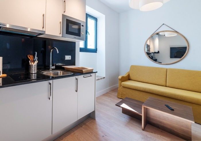 Cocina americana moderna y sala de estar con armarios blancos, sofá amarillo, espejo redondo y mesa de centro.