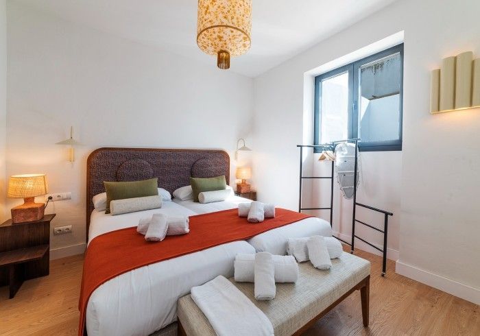 Habitación luminosa con una colcha roja, dos taburetes y una ventana con cortinas blancas.