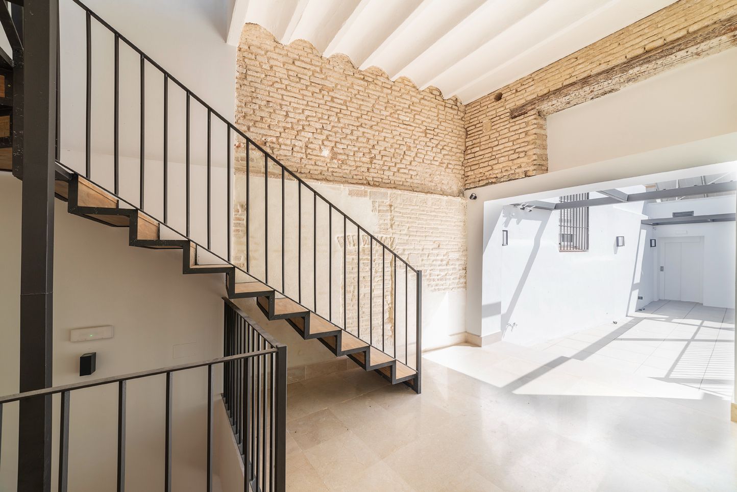 Escalera luminosa con barandilla de metal negro, pared decorativa de piedra y patio blanco bañado por el sol.