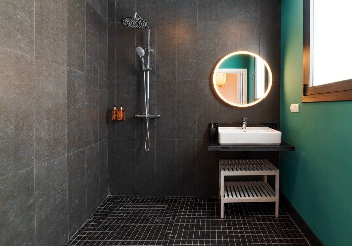 Baño moderno con azulejos oscuros, pared color turquesa, espejo redondo, ducha y lavabo blanco.