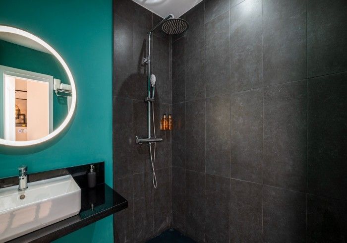 Baño moderno con pared color turquesa, espejo redondo, lavabo y azulejos negros en la ducha.