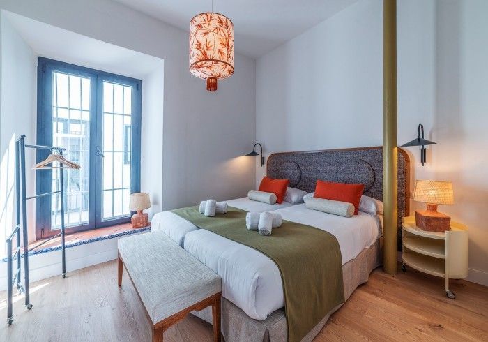 Habitación luminosa con cama, ventana azul, cabecero gris y cálido suelo de madera, iluminada por una lámpara colgante.
