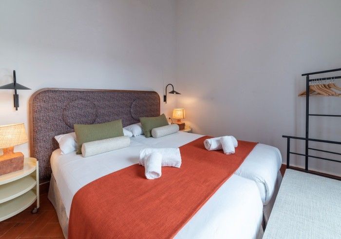 Acogedora habitación con alfombra naranja, ropa de cama blanca, lámpara de mimbre y perchero negro.