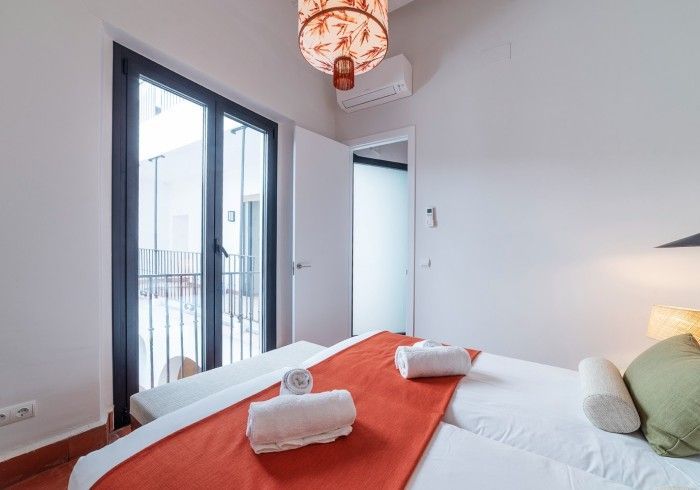 Habitación luminosa con colcha naranja, toallas blancas, puertas que dan al balcón y lámpara colgante tipo farol.