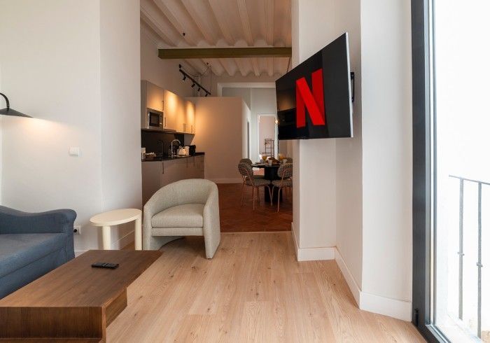 Salón moderno con suelo de madera clara, sillas blancas y un televisor montado en la pared.