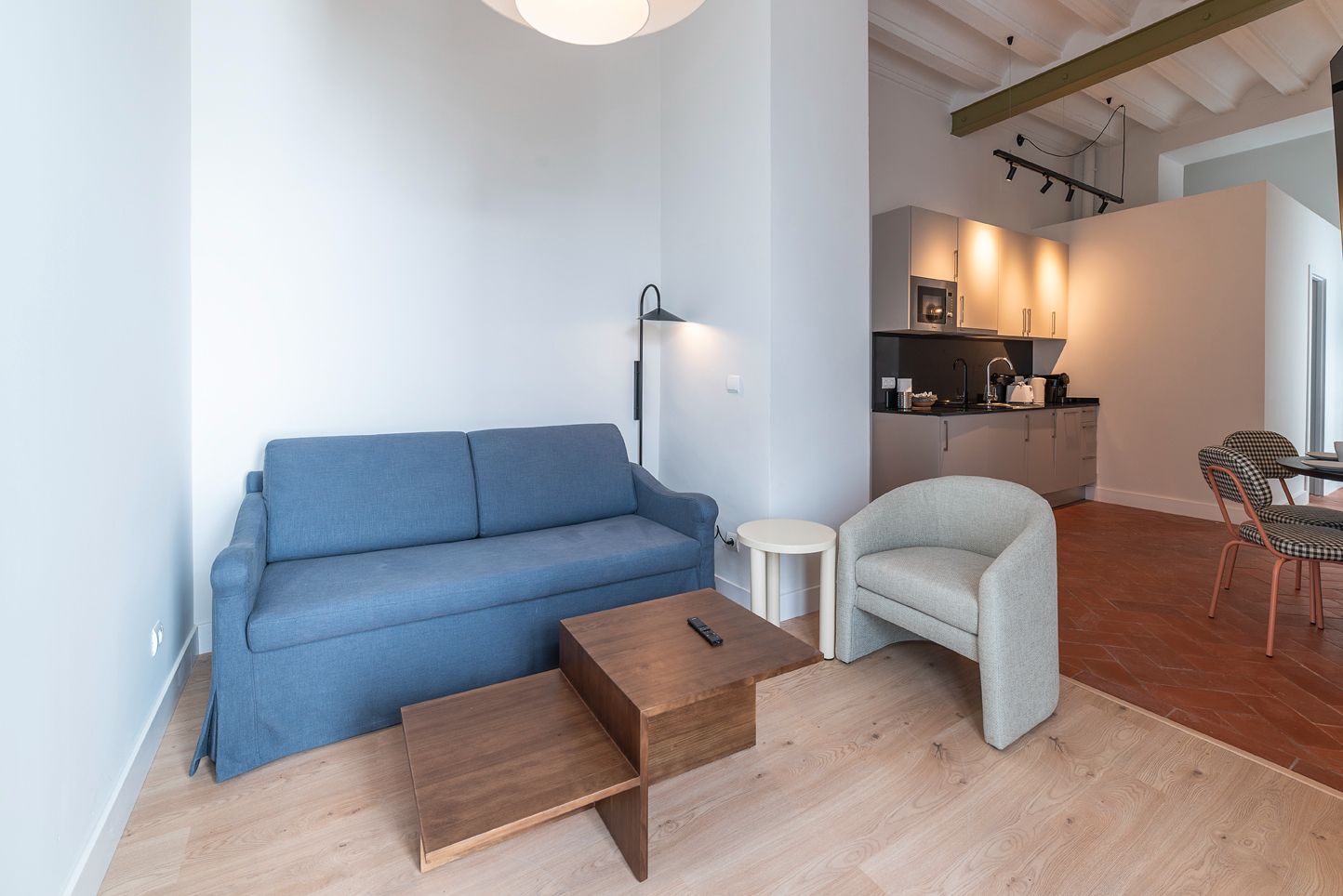 Sala de estar moderna con sofá azul, silla blanca, mesas de madera y área de cocina abierta.