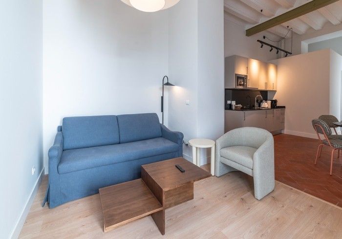 Sala de estar moderna con sofá azul, silla beige, mesa de centro de madera y área de cocina abierta.