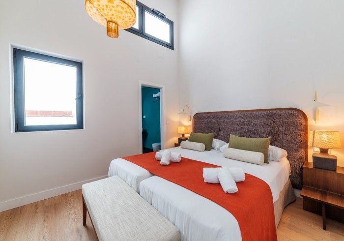 Dormitorio moderno con una cama grande, camino de mesa naranja, cabecero tejido y dos ventanas con lámparas colgantes.