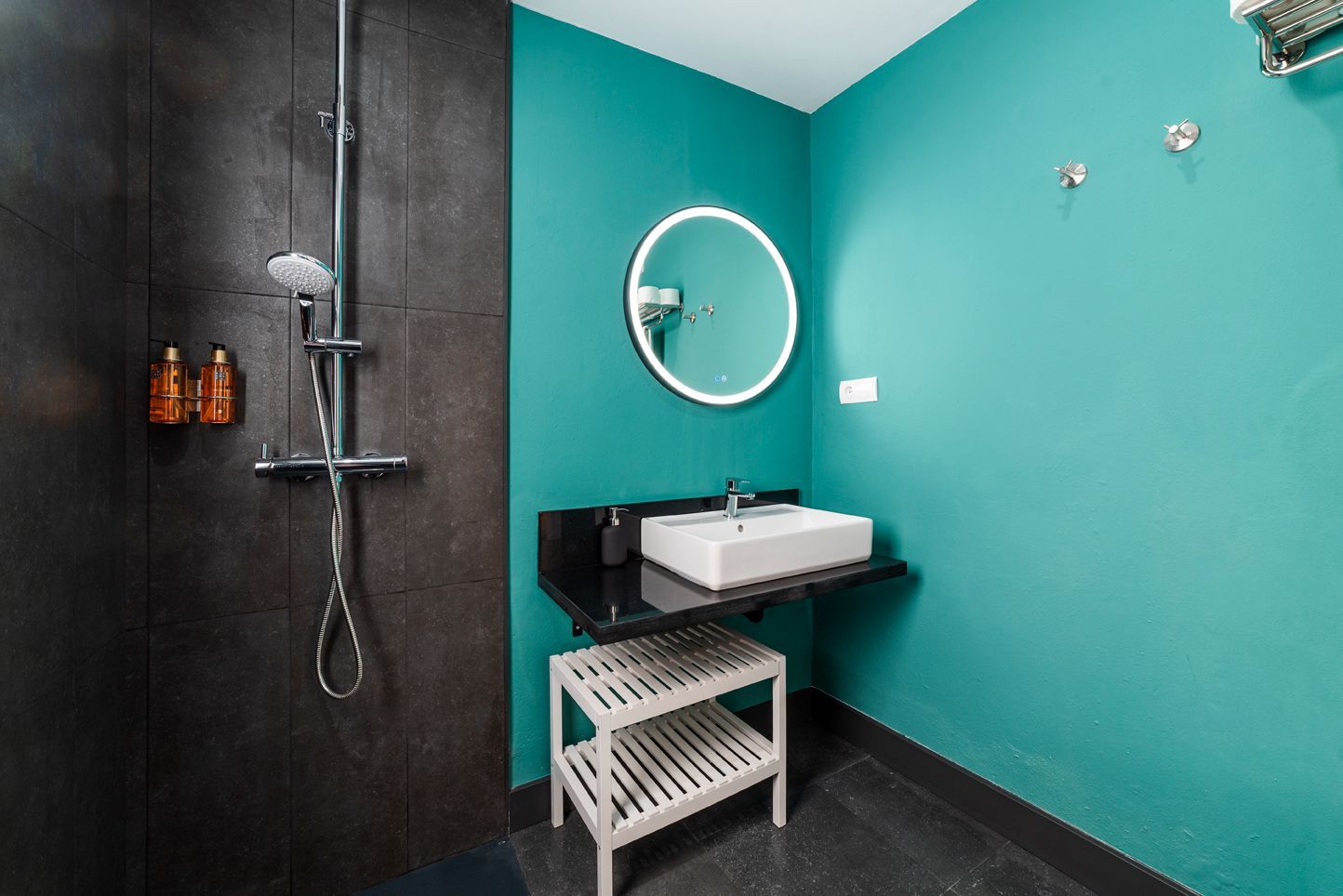 Baño moderno con pared de ducha negra, pared color turquesa, espejo redondo, lavabo y banco blanco debajo del tocador.