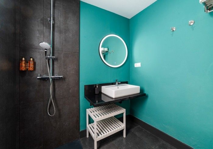 Baño moderno con paredes color turquesa, azulejos negros, ducha a ras de suelo, lavabo y espejo redondo.