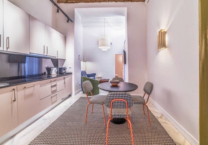 Cocina-comedor moderna y estrecha con paredes blancas, armarios de madera clara, mesa redonda y sillas negras.
