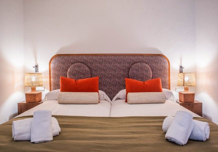 Cama bien hecha con cabecero tejido, cojines decorativos naranjas y lámparas de noche en un dormitorio luminoso.
