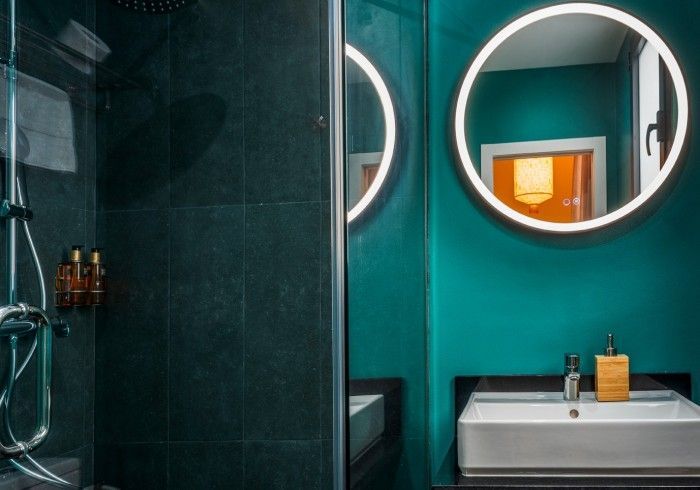 Baño moderno con paredes color turquesa, espejo redondo iluminado y lavabo blanco junto a una mampara de ducha de cristal.