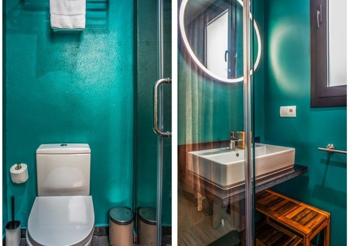 Baño color turquesa con inodoro, ducha de cristal y lavabo mural junto a un banco de madera.