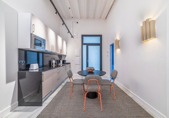 Cocina moderna y rincón de comedor con paredes blancas, armarios grises, ventanas con marcos azules y una mesa redonda.