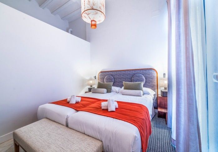 Habitación luminosa y moderna con paredes blancas, una cama con una manta roja y cortinas junto a la ventana.