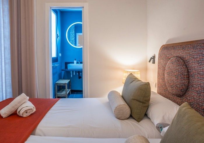 Habitación de hotel con dos camas, sábanas blancas, manta roja y puerta de baño azul abierta.