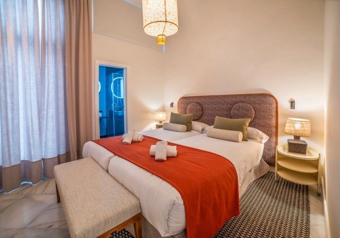 Acogedora habitación de hotel con cama, manta naranja, lámparas de noche y cortinas transparentes.