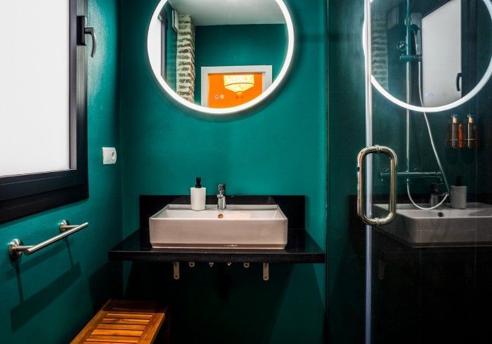 Baño moderno con paredes color turquesa, espejo redondo, lavabo blanco y mampara de ducha de cristal.