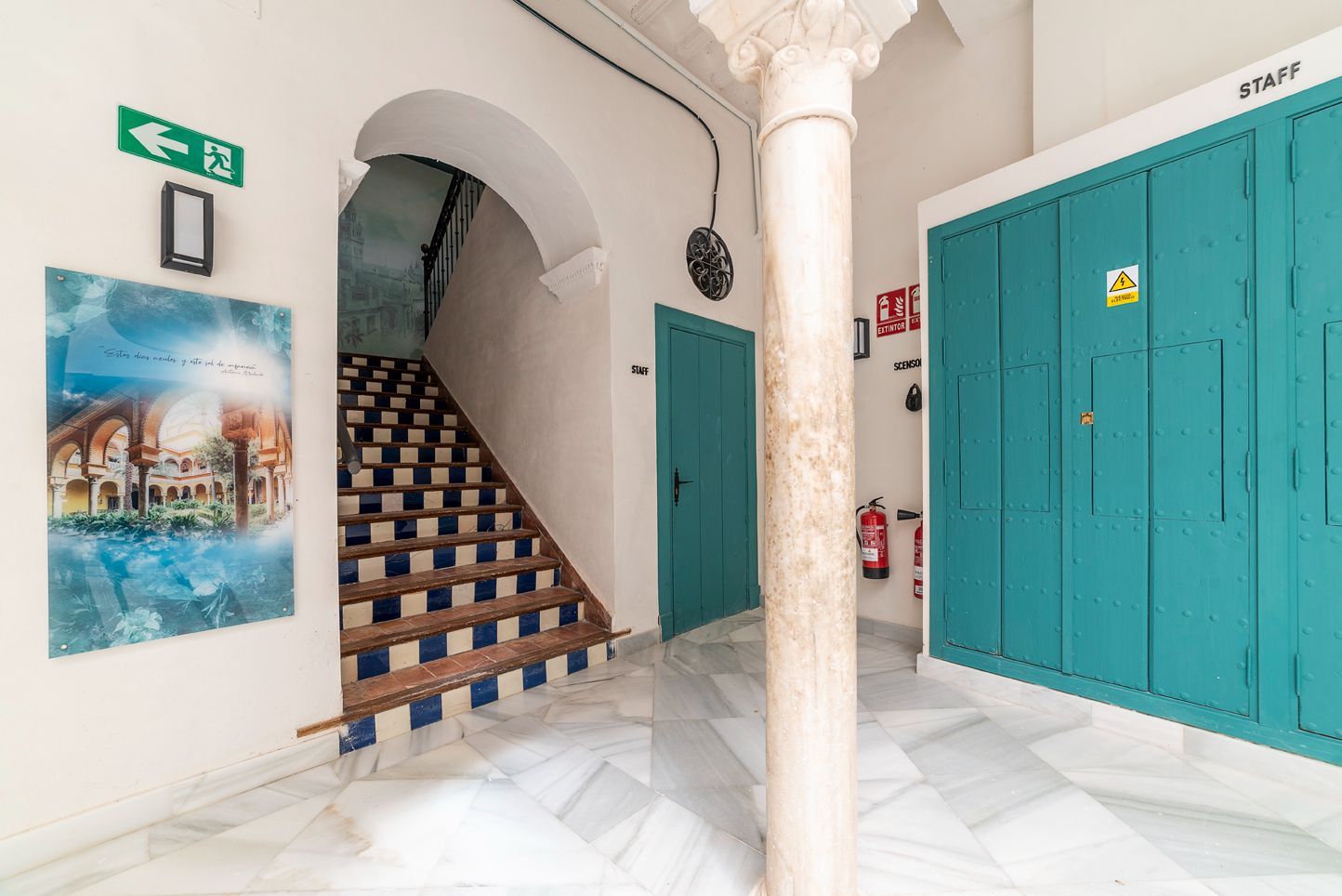 Pasillo con azulejos blancos, puertas turquesas, escalera arqueada y una columna central.