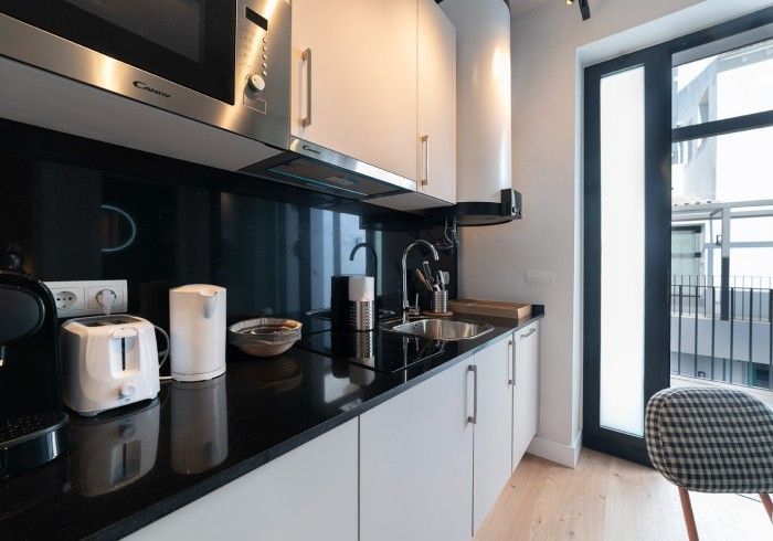 Cocina moderna con encimera negra, armarios blancos, pequeños electrodomésticos y puerta de cristal que da a un balcón.