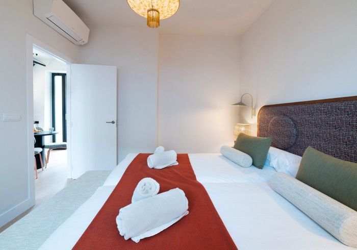 Habitación luminosa y moderna con cama blanca, alfombra roja, almohadas verdes y puerta abierta a una zona de estar.