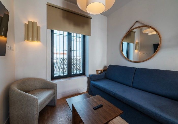 Acogedora sala de estar con sofá azul, silla gris, mesa de centro de madera y ventana con rejas.