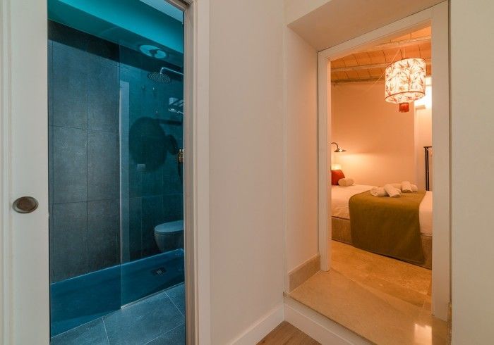Puerta del baño junto a un dormitorio con iluminación cálida, con una colcha verde y una lámpara colgante.