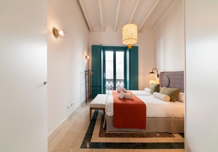 Habitación luminosa con cama, manta naranja, alfombra estampada, puertas color turquesa y lámpara colgante.