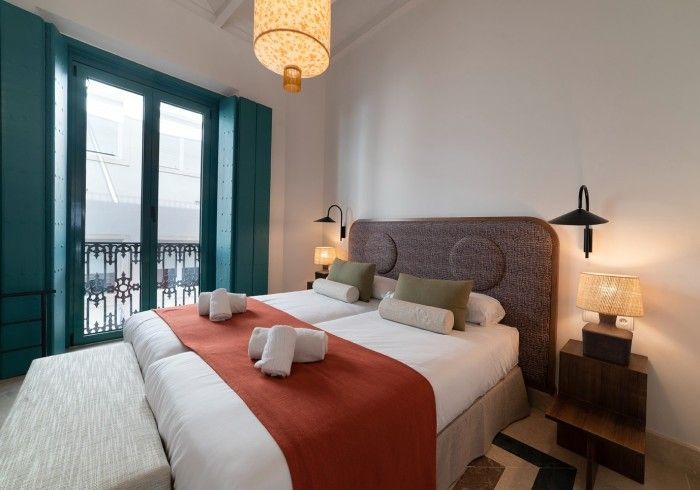 Habitación luminosa con una cama grande, puertas de balcón color turquesa y lámparas de noche cálidas.