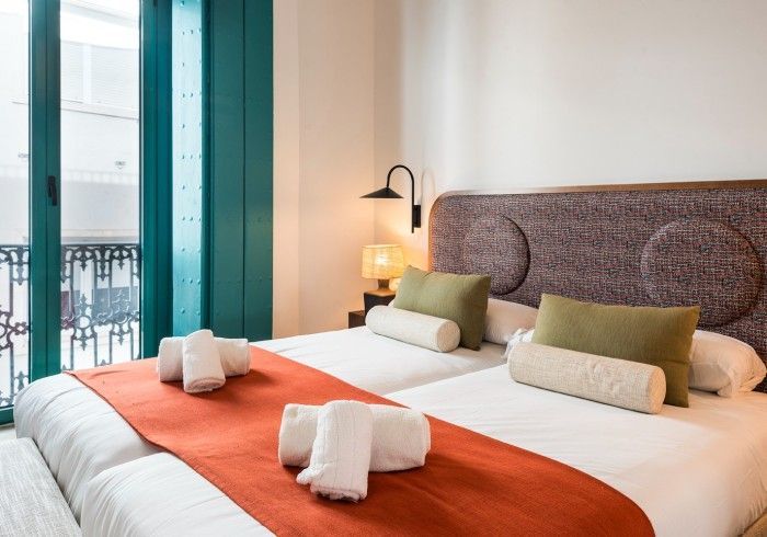 Habitación moderna con una cama grande, camino de mesa naranja, almohadas verdes y vista al balcón.