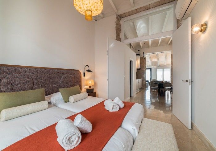Habitación luminosa con dos camas individuales, manta naranja, toallas blancas y vistas a un comedor.