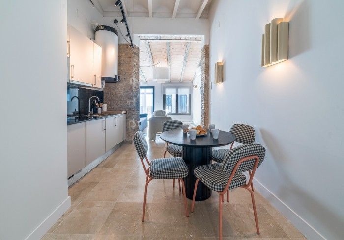 Comedor moderno y luminoso con mesa redonda negra, sillas estampadas y una pequeña cocina junto a la pared izquierda.