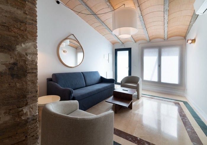 Sala de estar moderna con sofá azul marino, sillas blancas, espejo redondo y ventanas luminosas.