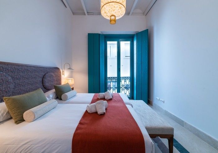 Habitación luminosa con cama blanca, alfombra roja, contraventanas color turquesa y puertas de balcón que dejan pasar la luz.