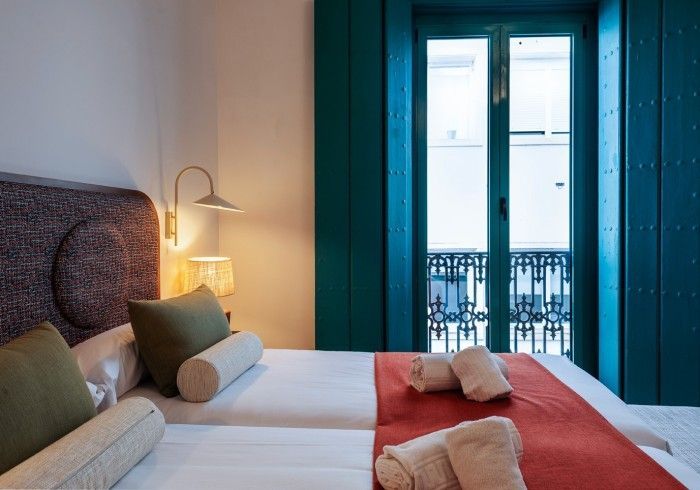 Acogedora habitación de hotel con dos camas individuales, luz cálida de lámpara, contraventanas color turquesa y balcón.