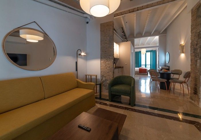 Elegante salón con sofá color mostaza, silla verde, espejo redondo y zona de comedor en el luminoso pasillo del apartamento.
