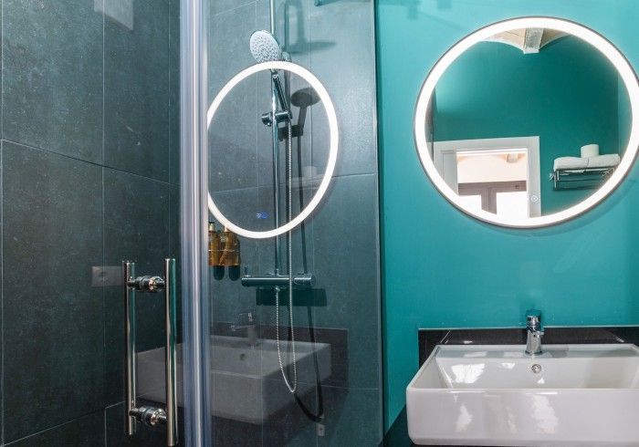 Baño moderno con ducha de cristal, espejo redondo iluminado y pared color turquesa con lavabo.