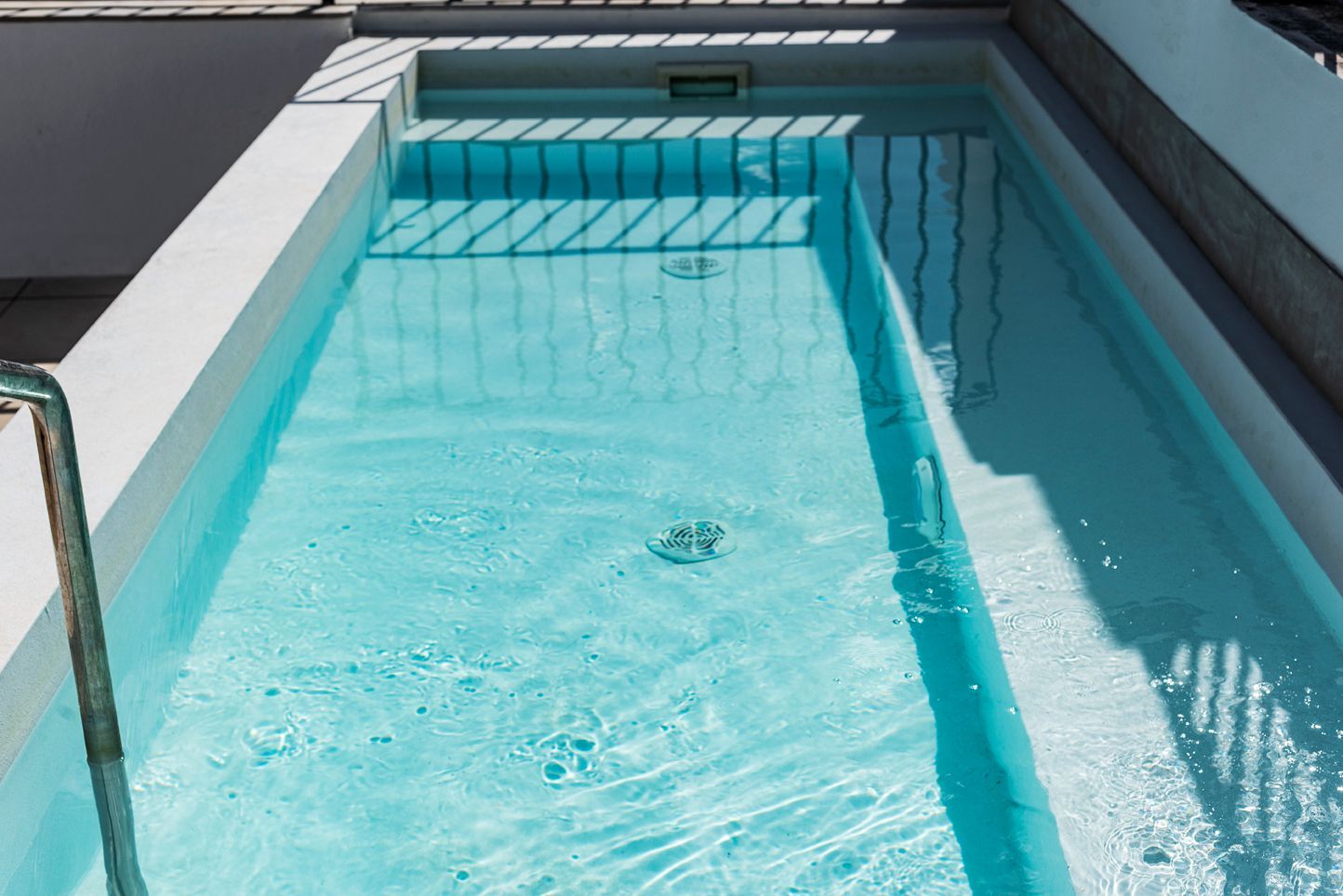 Pequeña piscina turquesa con agua cristalina, bordes de hormigón y barandilla metálica, bañada por la luz del sol.