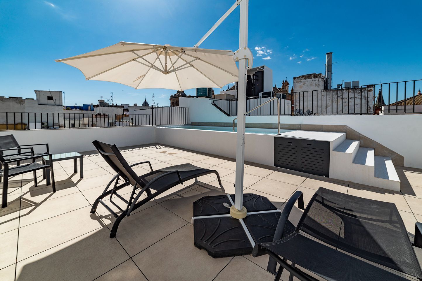 Terraza en la azotea con tumbonas, una gran sombrilla blanca y vistas panorámicas de la ciudad bajo un cielo azul brillante.
