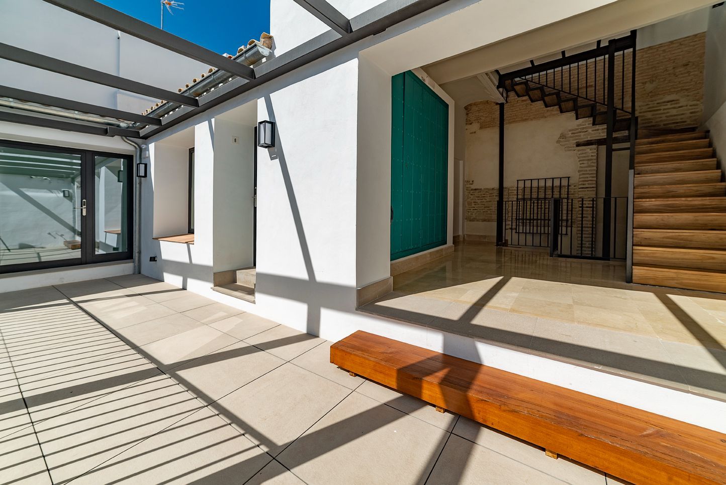 Patio soleado con sombra de pérgola, puertas de cristal abiertas y una escalera de madera que conduce al interior.