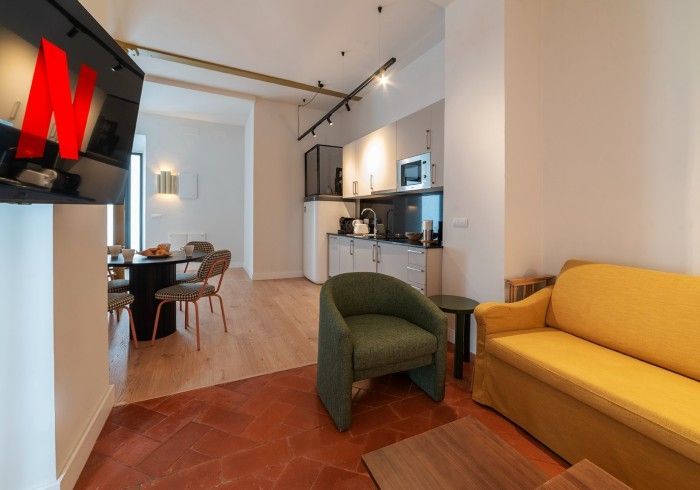 Sala de estar luminosa con sofá amarillo, silla verde, zona de comedor y cocina abierta al fondo.