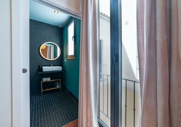 Baño compacto de color verde azulado con espejo redondo, lavabo y suelo estampado, visible a través de puertas correderas.