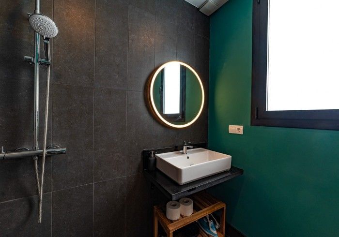 Baño moderno con paredes de azulejos oscuros, pared decorativa de color verde azulado, espejo redondo, lavabo, ventana.